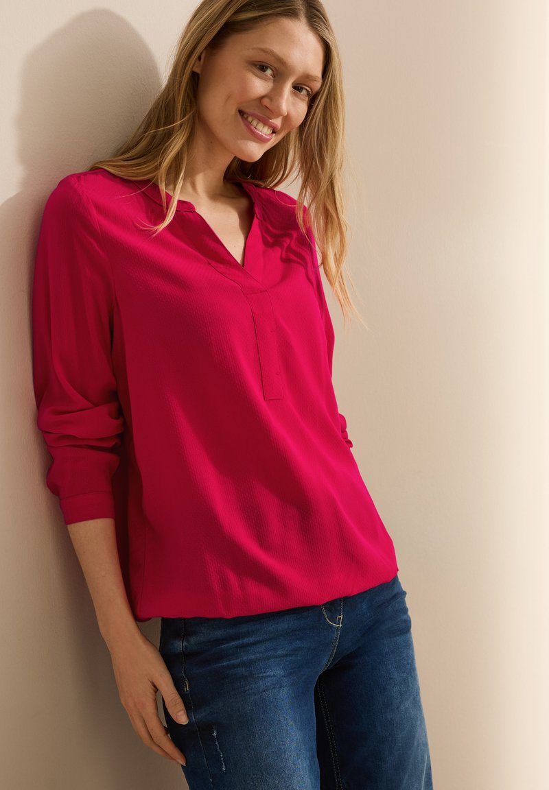 Blouse fuchsia avec une texture subtile, décolleté en V et manches retroussées, associée à un jean en denim bleu foncé.