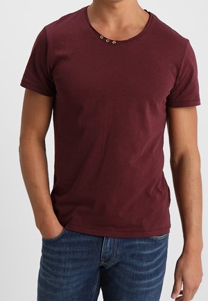 Bordeauxrotes T-Shirt mit kurzen Ärmeln, rundem Halsausschnitt und drei braunen Knöpfen, die den Kragen betonen, kombiniert mit Jeansstoff-Jeans.