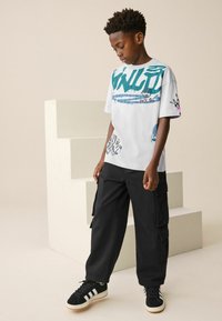 Weißes Kurzarm-T-Shirt mit türkisem Grafikdesign, kombiniert mit schwarzen Cargo-Hosen und schwarzen Sneakers mit weißen Streifen.