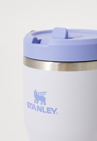 White stainless steel travel mug with a light purple plastic lid and a silver band. Features a blue logo of a lion with wings, "STANLEY".