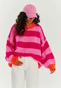 Pestrý pruhovaný oversized svetr v odstínech růžové a oranžové s hrubým pleteným vzorem. Spárován s odpovídající růžovou čepicí a bílými kalhotami.
