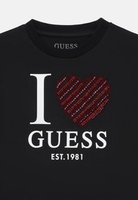 Juodas marškinėliai su balta "I" ir "GUESS EST. 1981" tekstu. Išsiskiria raudonu širdies motyvu, pagamintu iš dryžuotų akmenukų, kuri suteikia tekstūrinį akcentą.