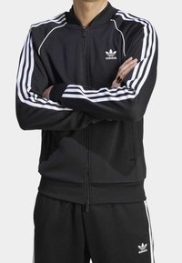 Svart zip-up träningsjacka med vita tre-stripes detaljer på ärmarna, ribbade mudd och en rund krage. Har en Adidas-logga på bröstet.
