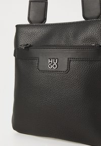 Črna teksturirana usnjena torbica z zadnjim žepom na zadrgo in vtisnjenim logotipom "HUGO". Torbica ima dve kratki ročici.