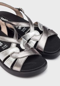 Sandalias metálicas plateadas con acabado texturizado, que presentan tiras entrelazadas y una suela negra acolchada. Incluye correa ajustable para el tobillo.