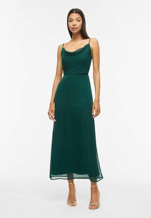 Femme debout portant une robe maxi vert foncé sans manches avec de fines bretelles et des sandales à talons dorées sur un fond blanc uni.