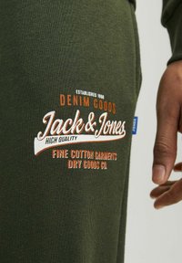 Grøn bomuldsbeklædning med en struktureret overflade. Frontdesignet inkluderer hvide og orange broderede brandingelementer.