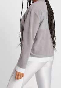 Grå fleece pullover med lange ærmer og en lysegrå panel på den øvre ryg. Har en blank sølvkant og manchetter. Båret med blanke leggings.
