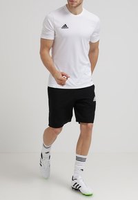 Bílé sportovní tričko s krátkým rukávem a černým logem Adidas, kombinované s černými šortkami. Také obsahuje bílé ponožky a fotbalové boty s černými akcenty.