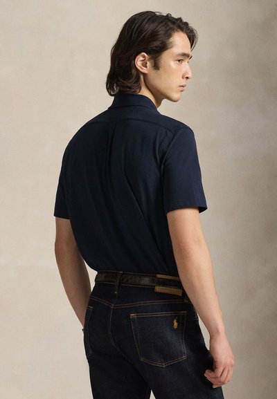 Camicia blu navy a maniche corte in tessuto liscio, con colletto e giogaia posteriore. Abbinata a jeans di denim scuro e cintura marrone.