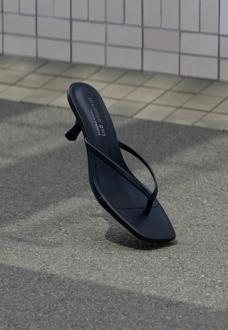 Μαύρο πέδιλο με ψηλό τακούνι, τετράγωνη μύτη και λεπτά λουριά, όρθιο σε γκρι πλακόστρωτο κοντά σε τοίχο με πλακάκια.