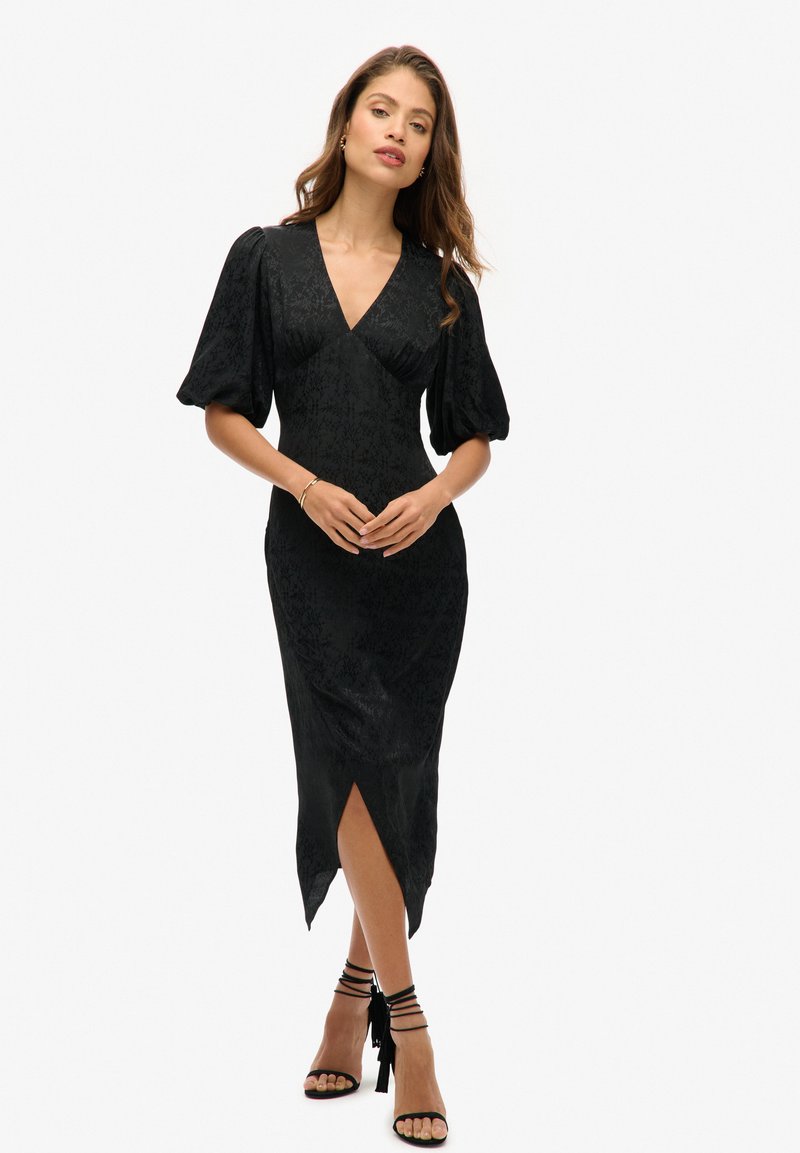 Vestido midi negro con mangas abullonadas, escote en V y textura floral. Presenta una abertura alta en el muslo y está estilizado con sandalias negras con tiras.