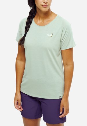 Femme aux cheveux tressés portant une chemise à manches courtes vert clair et un short violet, debout devant un fond uni.