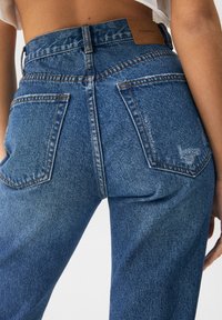 Jeans de mezclilla en azul medio, con una cintura alta, cinco bolsillos y un sutil desgaste en la parte posterior, con una etiqueta de marca marrón en la cintura.