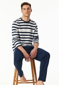 Gestreiftes Langarmoberteil aus navy- und weißen Baumwolle mit Rundhalsausschnitt und gesticktem Logo, kombiniert mit navy Hosen auf einem Holzstuhl.