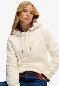 Felpa con cappuccio color avorio con finitura testurizzata, dotata di tasca centrale, coulisse e ricamo sottile. Indossata sopra una camicia a quadri.