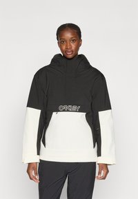 Czarny i biały jacket Oakley z wysokim kołnierzem, zapinany na zamek, z bocznymi kieszeniami i logo na piersi. Wykonany z trwałego, odpornego na warunki atmosferyczne materiału.