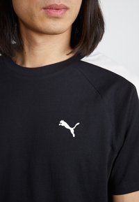 Juoda medvilninė marškinėliai su baltu Puma logotipu kairėje krūtinės pusėje ir kontrastingomis baltomis raglan rankovėmis. Lygi tekstūra ir standartinė sukirpimas.