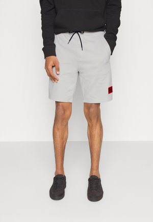 Mann trägt hellgraue Shorts mit Kordelzug und rotem Fleck am linken Bein, schwarzen Hoodie und schwarze Sneakers, steht mit teilweise in den Taschen steckenden Händen.