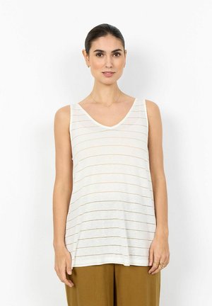 Femme aux cheveux foncés attachés, portant un haut blanc sans manches à encolure en V avec de fines rayures horizontales, et un pantalon marron, debout devant un fond blanc.