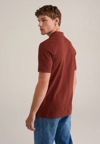 Bordeaux kortærmet polo-shirt med krave, parret med lyseblå jeans. Skjorten har sideslids og et glat, tekstureret stof.