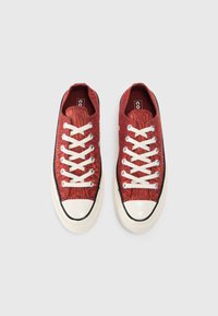 Røde teksturerte lerretssneakers med hvite snørebånd, svart gummitåhette og kremfarget såle. Har sirkulære lufthull og kontrasterende svart kant.