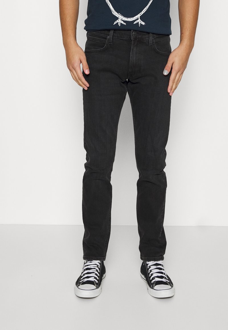 Personne portant un jean slim noir, des baskets noires et blanches, et une chemise bleu foncé avec un dessin de collier blanc, debout sur un fond blanc.