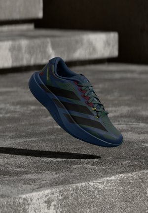 Ciemnoniebieski i czarny but do biegania z żółtymi akcentami unoszący się nad betonowymi schodami rzucającymi cień na ziemię.