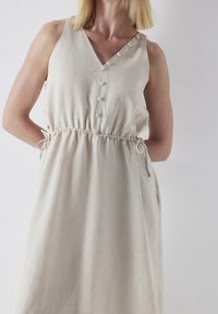 Robe en lin beige avec un col en V, dotée de boutons le long de l'épaule et d'une taille élastique. Poches intégrées au design.