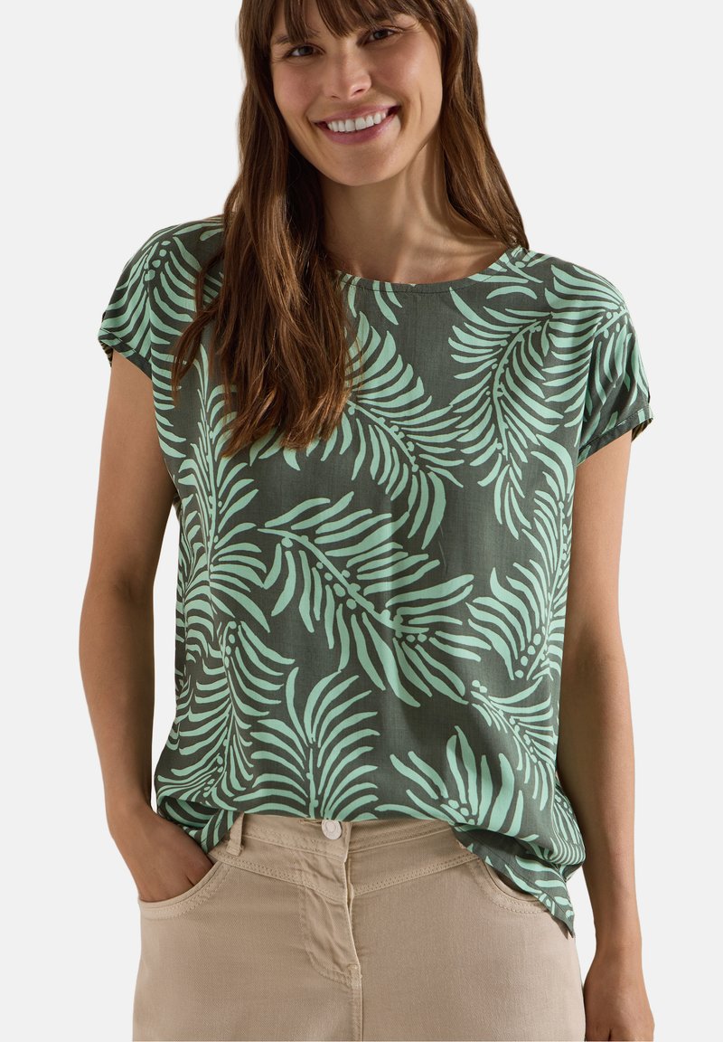 Femme souriante portant une chemise verte à manches courtes avec des motifs de feuilles vert clair et un pantalon beige, mains dans les poches, sur un fond uni.