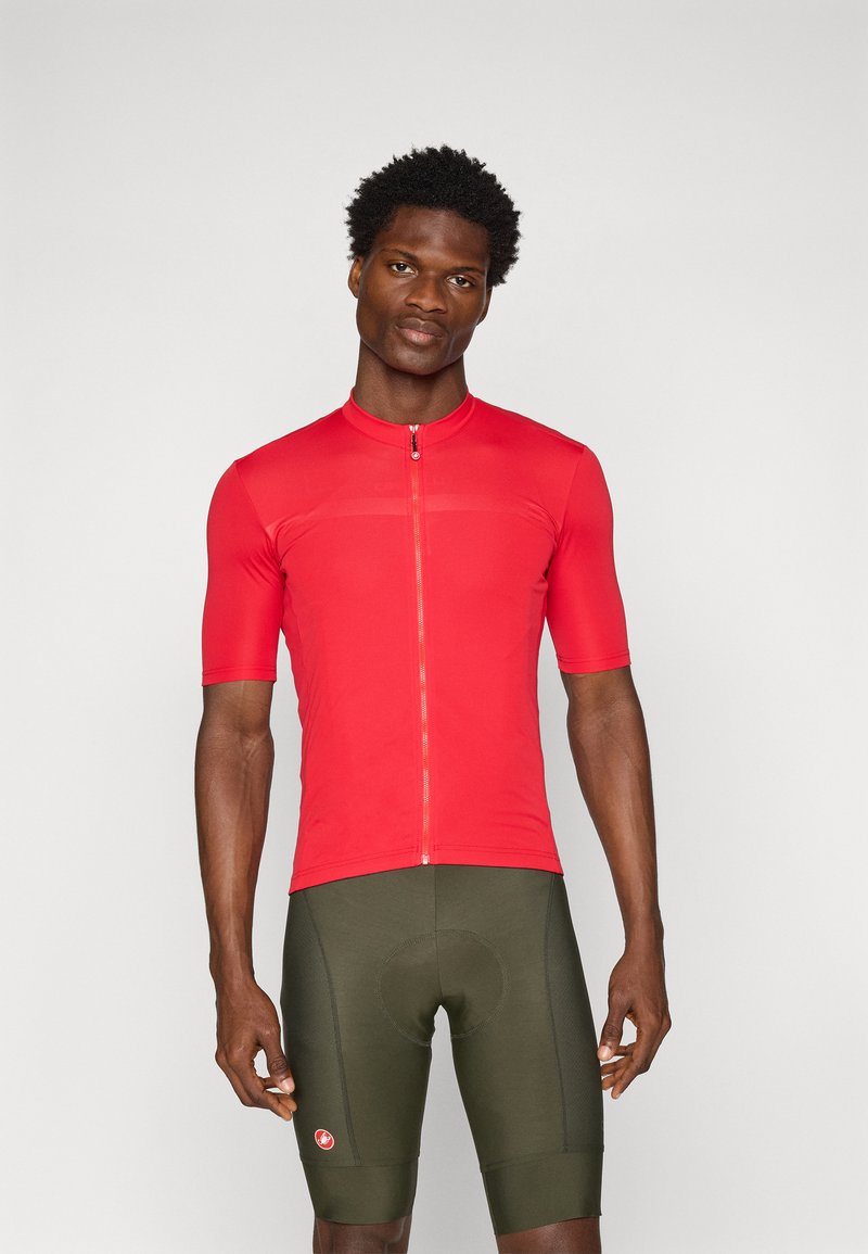 Maillot de cyclisme rouge à manches courtes avec une demi-fermeture éclair, fabriqué à partir d'un tissu élégant, porté avec un short rembourré vert olive arborant un écusson.