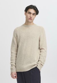 Suéter de punto beige con cuello redondo, puños y dobladillo acanalados; ajuste holgado con una apariencia suave y texturizada. Llevado con pantalones oscuros.