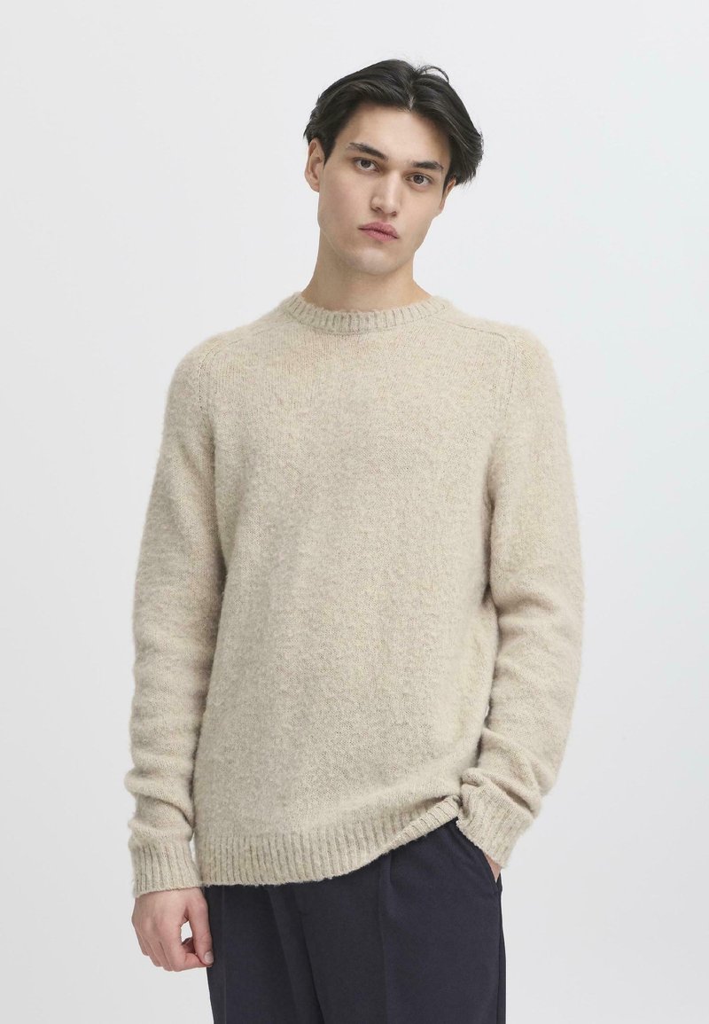 Suéter de punto beige con cuello redondo, puños y dobladillo acanalados; ajuste holgado con una apariencia suave y texturizada. Llevado con pantalones oscuros.