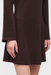 Gros plan d'une personne portant une robe en maille marron foncé à manches longues, les mains détendues sur les côtés et les jambes nues visibles sous l'ourlet.
