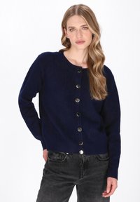 Tmavě modrý cardigan vyrobený z měkké tkaniny, s kulatým výstřihem a šesti kovovými knoflíky vpředu. Úzký střih s dlouhými rukávy.