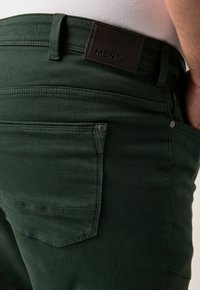 Dettaglio ravvicinato di jeans da uomo verde scuro che mostra la tasca posteriore, l'asola della cintura e il patch del brand in pelle nera con il testo "MEN+".