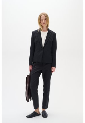 Black tailored blazer and matching trousers with a white top, paired with black slip-on shoes and a brown leather tote bag.