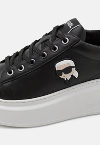 Sorte lærersneakers med en tykk hvit såle, med svarte snøresko, sølv øyelets og en tegneserie-logo detalj på siden.