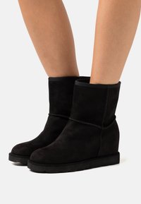Botines de ante negros con tacón cuña, que presentan un detalle de costura y un ribete superior elástico para un ajuste ceñido. Textura suave.