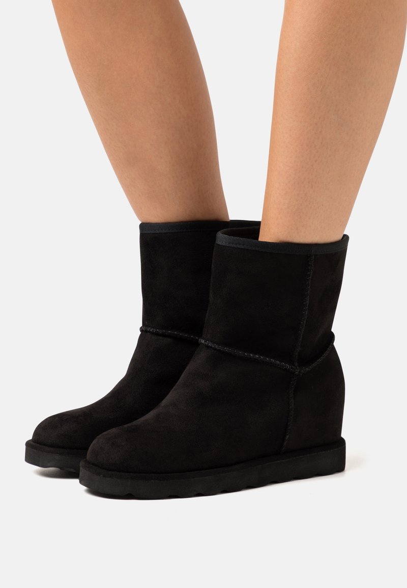 Botines de ante negros con tacón cuña, que presentan un detalle de costura y un ribete superior elástico para un ajuste ceñido. Textura suave.