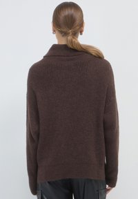 Donna con coda di cavallo castana, che indossa un maglione a coste marrone scuro e pantaloni neri, vista da dietro su uno sfondo neutro.