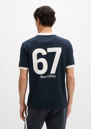 Homme de dos portant une chemise bleu marine à manches courtes avec col blanc, bordure blanche aux manches, grand "67" et "Marc O'Polo" dans le dos.