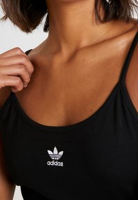 Svart ärmlös tanktop i slät tyg med en liten vit Adidas-logotyp på bröstet. Justerbara axelband.