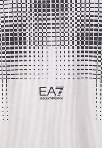 Balts sporta krekls ar melnu režģa rakstu, ribotu tekstūru un EA7 Emporio Armani logo melnā krāsā apakšējā labajā stūrī.