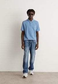 Camisola polo azul clara, jeans de lavagem clara e ténis brancos. Modelo usa óculos de sol escuros, de pé contra um fundo simples.