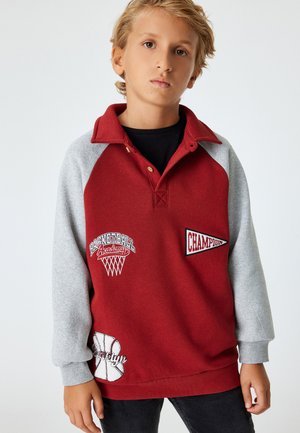 Pull rouge et gris avec un col, présentant des graphiques de basketball brodés et du texte. Tissu doux avec une coupe légèrement décontractée.