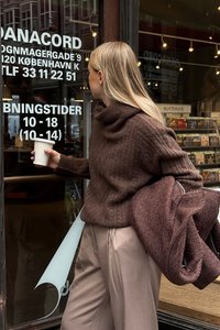 Ruskea neulepusero, jossa on kauluri ja teksturoitu kuvio, yhdistettynä vaaleanbeigeihin housuihin. Pitelee valkoista kahvikuppia ulkona kaupan edustalla.