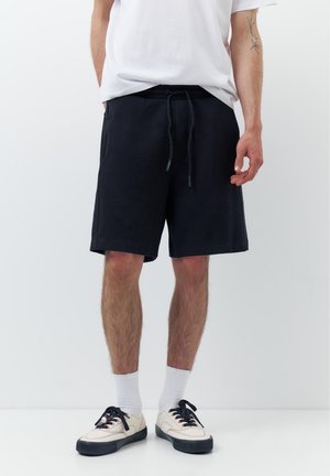Homme portant un short noir à cordon, des chaussettes blanches, des baskets blanches avec des lacets noirs et un t-shirt blanc, debout devant un fond uni.