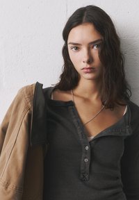 Dunkelgraues, geripptes Langarmshirt mit Knopfleiste, getragen unter einer hellbraunen Jacke. Sichtbare silberne Halskette. Weiche Textur, lässiges Design.