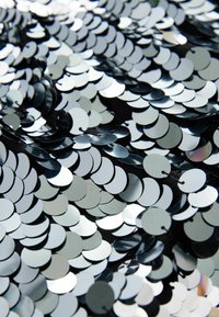 Silberne Pailletten dicht auf einem schwarzen Stoffhintergrund angeordnet, mit unterschiedlichen Oberflächen und reflektierenden Effekten, die ein strukturiertes Aussehen schaffen.